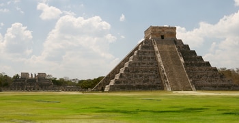 a large brick building with a grassy field with Chichen Itza in the background