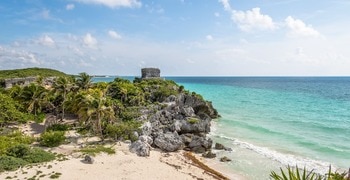 a beach with palm trees and a stone structure on the shore with Tulum in the background